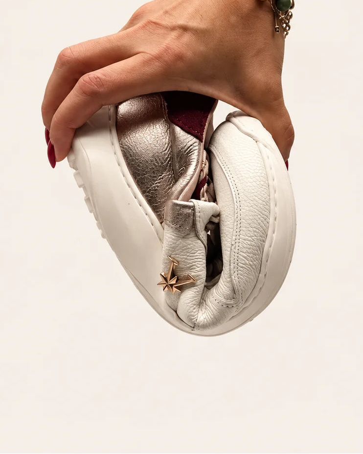 Mano doblando la deportiva barefoot de transición de mujer modelo LÍA nácar en piel color nata con detalle en platino y talón en burdeos de Valenta Unic para mostrar la flexibilidad de su suela